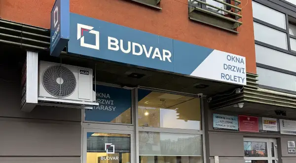 Szyld salonu okien i drzwi "BUDVAR" z logo, klimatyzatorem i napisem "OKNA DRZWI ROLETY".