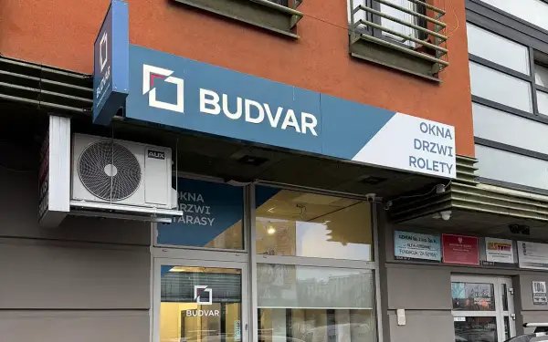 Szyld salonu okien i drzwi "BUDVAR" z logo, klimatyzatorem i napisem "OKNA DRZWI ROLETY".